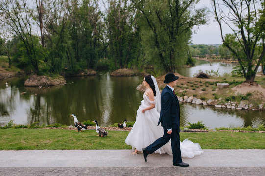 Beautiful newlyweds pass by each other and walk against the backdrop of nature and the lake. Wedding portrait of a stylish groom in a hat and a young bride with curly hair. Concept and photography.