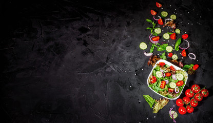Various fresh mix salad leaves with tomato and cucumber in a glass bowl on dark background with ingredients. Top view with copy space