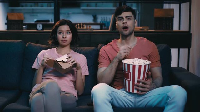 young, attentive man and woman eating snacks while watching tv on couch at home