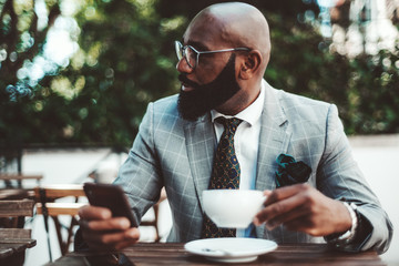 An elegant black bald senior with beard and eyeglasses and in a formal costume is having a coffee...