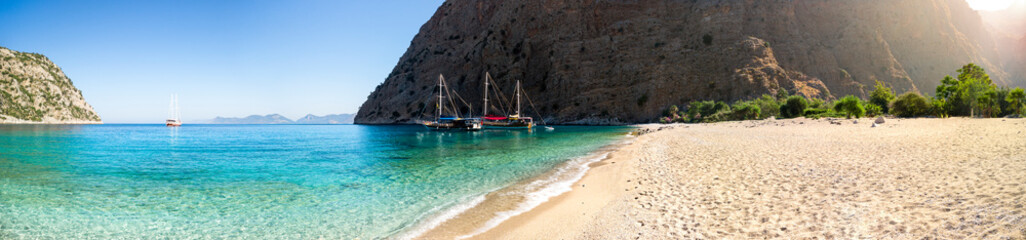 Fototapeta na wymiar Sailing ships moored in a secluded bay with turquoise water and empty beach at sunrise, Oludeniz, Turkey panoramic