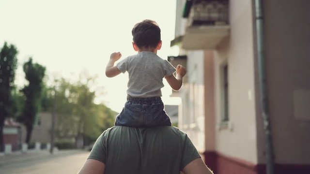 Father and son walking in city at sunset. Two year old kid is sitting on the parent's shoulders. Happy family concept. Footage in slow motion