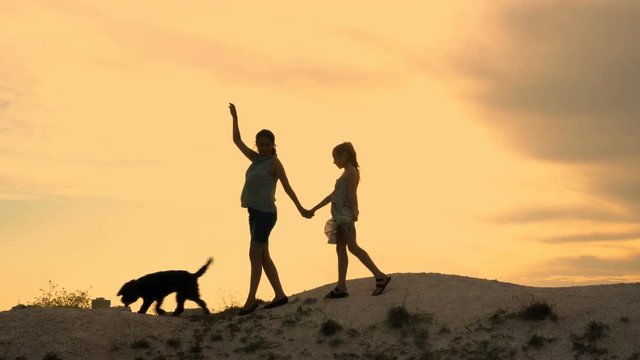 Happy family: mom, little girl and their dog stand on the top of the hill, they wave their hands goodbye and go down against the dramatic sky. Silhouette. General view. 4K