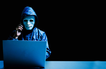 Anonymous computer hacker in white mask and hoodie. Obscured dark face using laptop computer for...