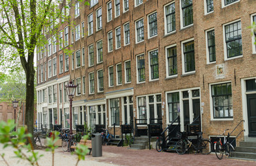 Amsterdam, Netherlands - May 03 2019. Street view of long brick house with apartments with big high windows and bicycles near entrance.