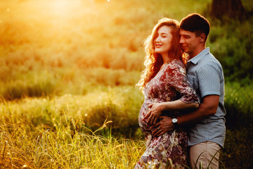 Happy young parents, mom and dad, hugging baby bump and smiling, enjoying beautiful moment at sunset among grass.