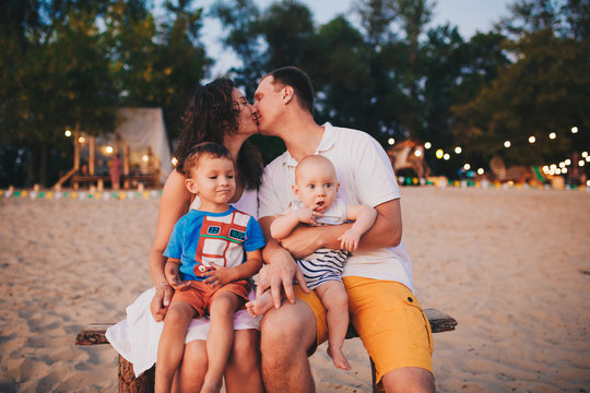 The concept of a family vacation. Young family sitting on a bench in the evening on a sandy beach. Mom and Dad kiss, the older brother kisses the younger on the cheek.