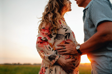 Stylish pregnant couple holding hands on belly in sunny light in autumn park.