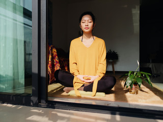 Pretty Chinese young woman meditating at home, sitting on floor with furry cushion in sun light,...