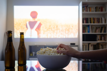 Home cinema entertainment: watching a film from a video projector in a room. Dim living room with a cinematic picture projected on the wall, human hand grabbing popcorn - obrazy, fototapety, plakaty
