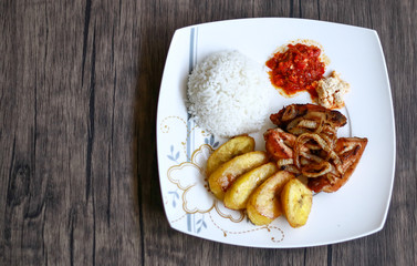 African fried chicken with fried bananas, rice, fried onions, chili sauce and candlenut sauce. Wood background.