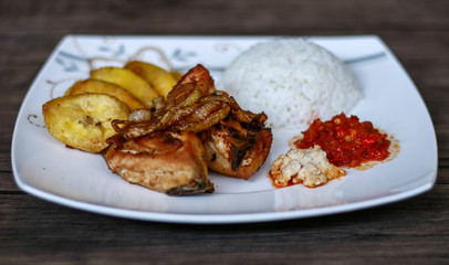 African fried chicken with fried bananas, rice, fried onions, chili sauce and candlenut sauce. Wood background.