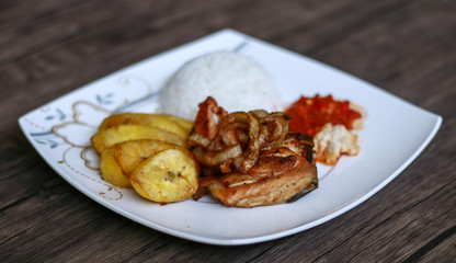 African fried chicken with fried bananas, rice, fried onions, chili sauce and candlenut sauce. Wood background.