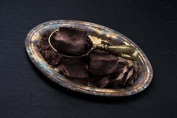 Chunks chocolate on metal tray and brass scoop on a black slate background