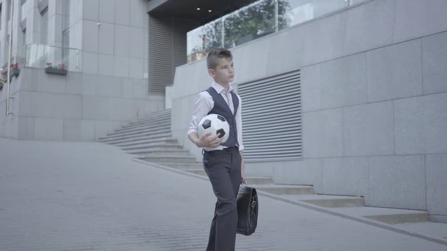 Serious Well-dressed Boy Walking Down The Street Holding The Soccer Ball And Purse In Hands. Serious Kid Simultaneously Acting Like Child And Adult. Bottom View