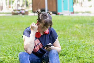Naklejka na ściany i meble Portrait of a teenage girl sitting on the grass in a city park in a blue t-shirt and jeans with red headphones.