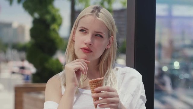 Attractive dreaming blonde girl thinking over something and coming up with an idea while eating an ice cream. Portrait of a creative girl holding an ice cone standing on the ciy street in sunny