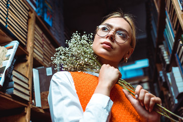 low angle view of woman in glasses holding white flowers and looking away in library