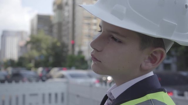 Cute little pensive boy wearing business suit and safety equipment and constructor helmet standing on a busy road in a big city. Engineer, architect, builder doing his work. Child as adult