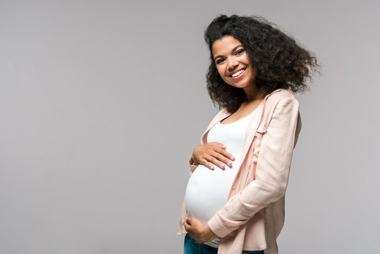 cheerful young pregnant african american woman touching belly on grey