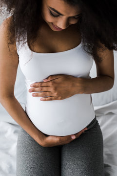 overhead view of positive curly african american pregnant woman touching belly while sitting on bed