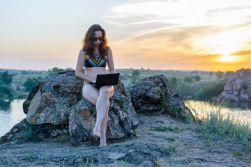 Young freelancer woman working on a laptop while sitting on a rock against the backdrop of a...
