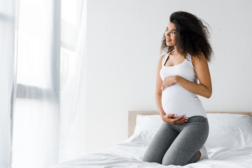 cheerful african american pregnant woman touching belly while sitting on bed