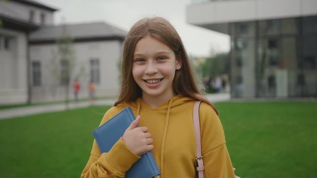 Joyful teenage student ready for school holding a book abd backpack staying in the school courtyard. Portrait of a pretty smart girl smiling outdoors near in the green area.