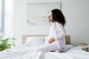 cheerful young pregnant african american woman sitting on bed and touching belly