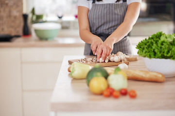 Dedicated Caucasian housewife in apron standing in kitchen and chopping mushrooms. On table are lots of vegetables. Cooking at home concept.
