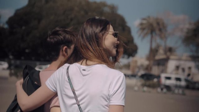 A back view of a young couple walking through a city a sunny day. The girls hand is on her boyfriends shoulder and his is on her back. There is a blurred summer view around them. With green trees