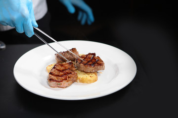 The cook puts tweezers on a plate of pieces of grilled meat with vegetables. The concept of cooking grilled meat indoors. High kitchen. Photo on a black background.