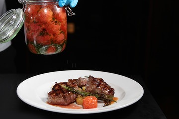 A cook decorates a cooked dish of grilled meat with vegetables canned salted tomato from a can. Hands cook, photo without a face. Macro photo. Concept of interior shooting.