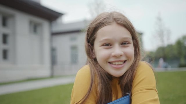 Portrait of a charming cute girl teenage student holding a book tightly smiling outdoors. Close-up of a happy and smart schoolgirl loves reading sitting in the open area near campus.
