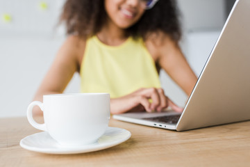 Fototapeta premium selective focus of cup with drink on wooden table near cheerful woman using laptop