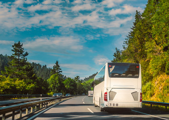 White tour bus or coach on a two lane motorway travelling through a tree lined hilly countryside on a sunny day with blue sky. © Christine Bird