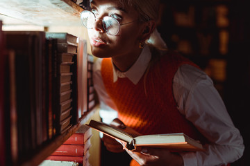 pretty woman in glasses holding book and looking away in library