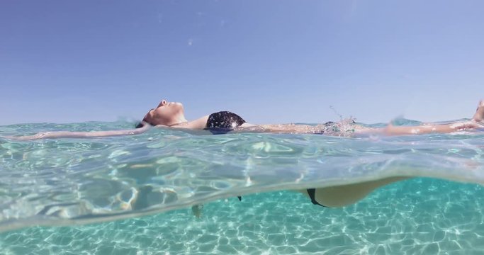 Slow motion of young woman is floating relaxed with belly up in a transparent clear blue water of tropical sea during her vacation.