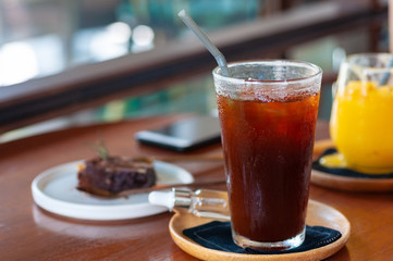 Fresh ice americano coffee with a piece of chocolate brownie in white plate on wooden table with double spoon and smart phone blur background.