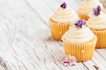 Beautiful vanilla cupcakes with buttercream icing decorated with sugar coated violet flowers. Selective focus with blurred background. - obrazy, fototapety, plakaty