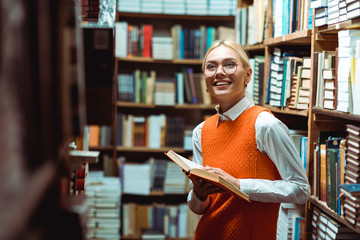 smiling and beautiful woman in orange dress holding book and looking away in library