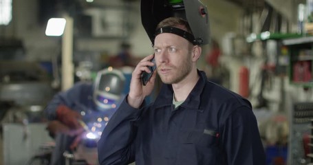 4K Mechanic wearing a welding helmet talking on phone in garage workshop