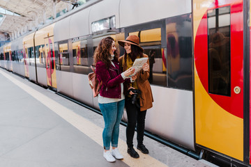 two women friends at train station waiting to take a train and travel. Reading a map and having fun. Tourism and lifestyle concept.