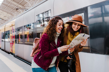two women friends at train station waiting to take a train and travel. Reading a map and having fun. Tourism and lifestyle concept.