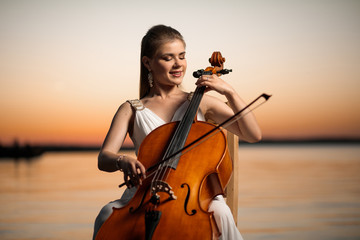  girl in a white dress playing the cello on the shore of the lake, after sunset.  © Ринат Куйшин