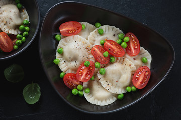 Black bowl of ravioli with cherry tomatoes and green peas, top view over black stone surface, studio shot
