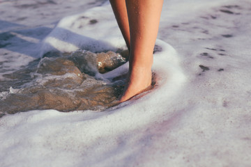 Female legs in the water. The girl goes along the coastline. Sunset light and sea foam.
