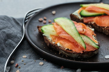 Close-up of open sandwiches with cream cheese, salmon and cucumbers on dark wooden plate on grey background covered dark grey napkin and pink sea salt with fork near the plate