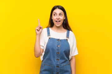 Young woman in dungarees over isolated yellow background intending to realizes the solution while lifting a finger up