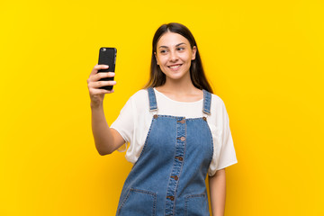 Young woman in dungarees over isolated yellow background making a selfie